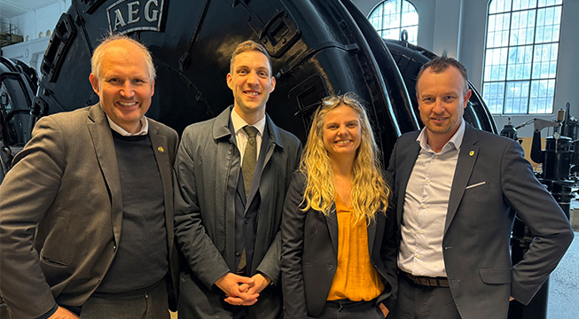 Bilde av tre menn i dress og en kvinne som står i en maskinhall på Vemork.