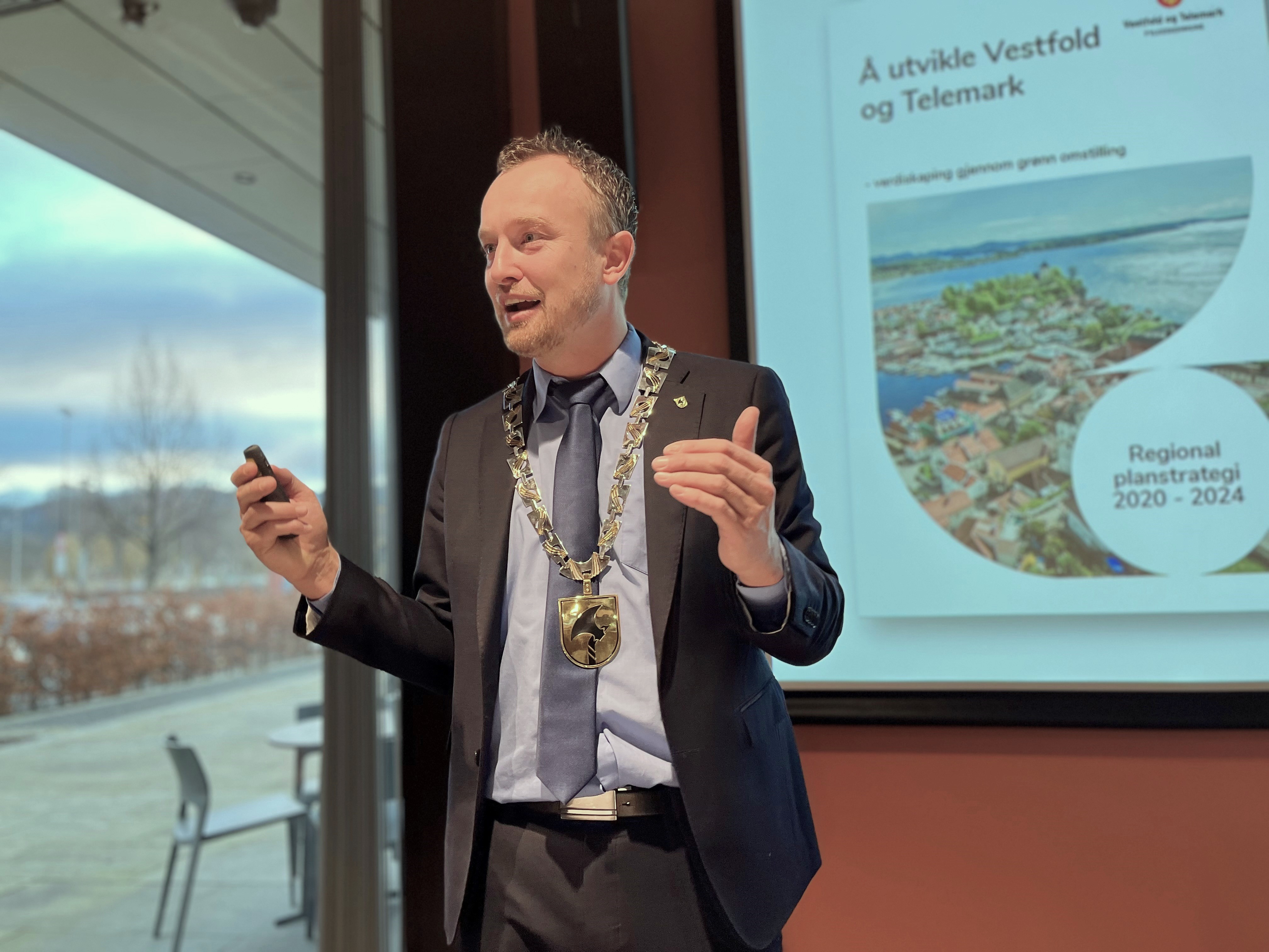 Mann i mørk dress og med et ordførerkjede rundt halsen som prater foran en skjerm med en presentasjon