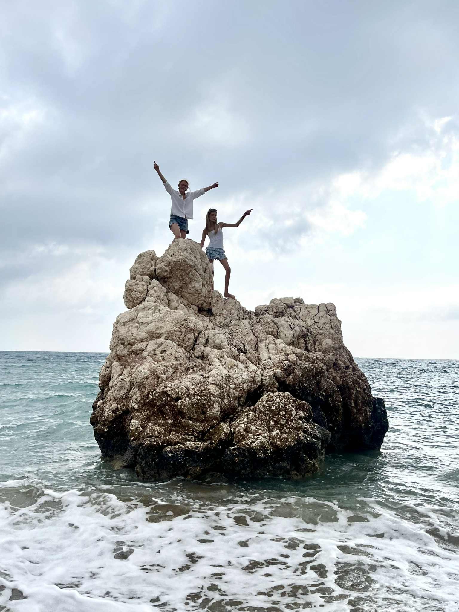 To jenter som står på en stor stein ute i havet. Begge jentene har armene opp mot himmelen.