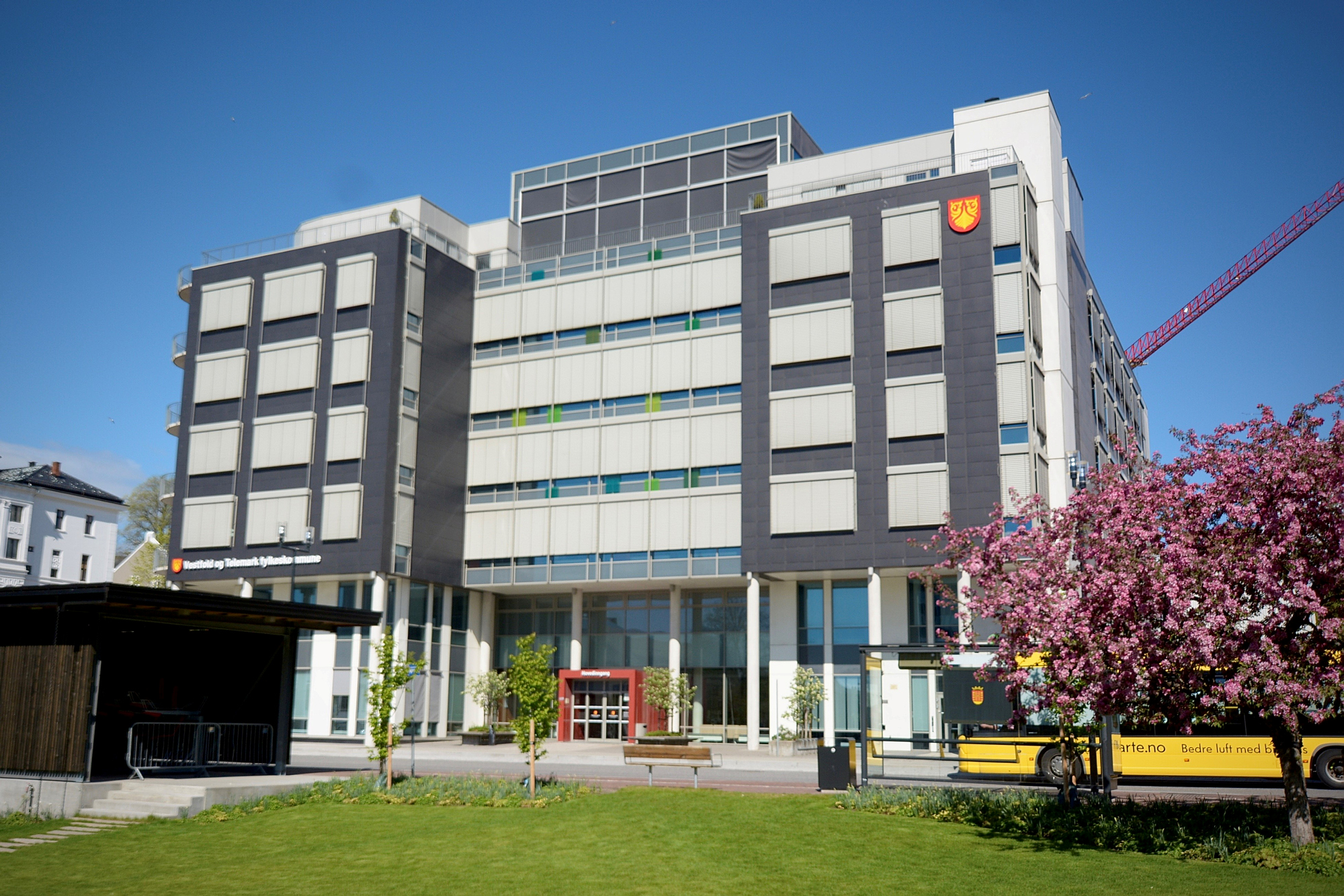 Stort bygg med en plen foran og en gul buss som passerer. Foto.