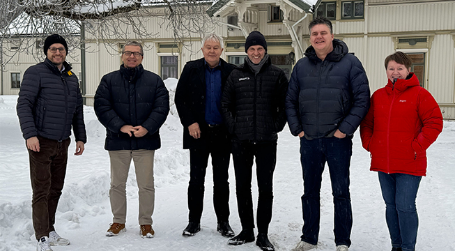 Fem menn og en kvinne står foran et eldre hus og smiler mot fotografen.