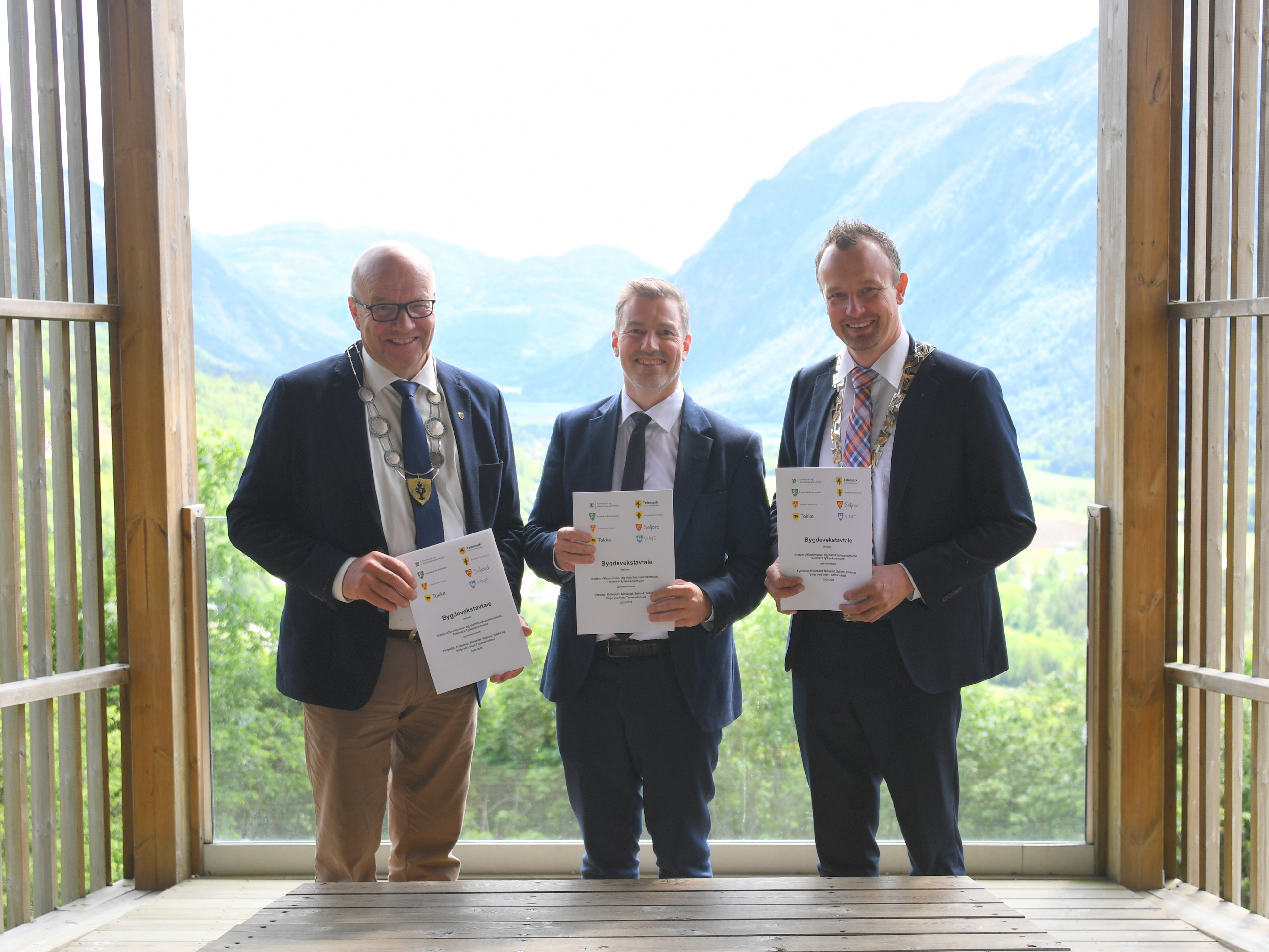 Fra venstre ordfører i Kviteseid og regionleder Tarjei Draugedal, statssekretær Emil Raaen og fylkesordfører Sven Tore Løkslid viser fram kontrakten med utsikten over Flatdal i bakgrunn.