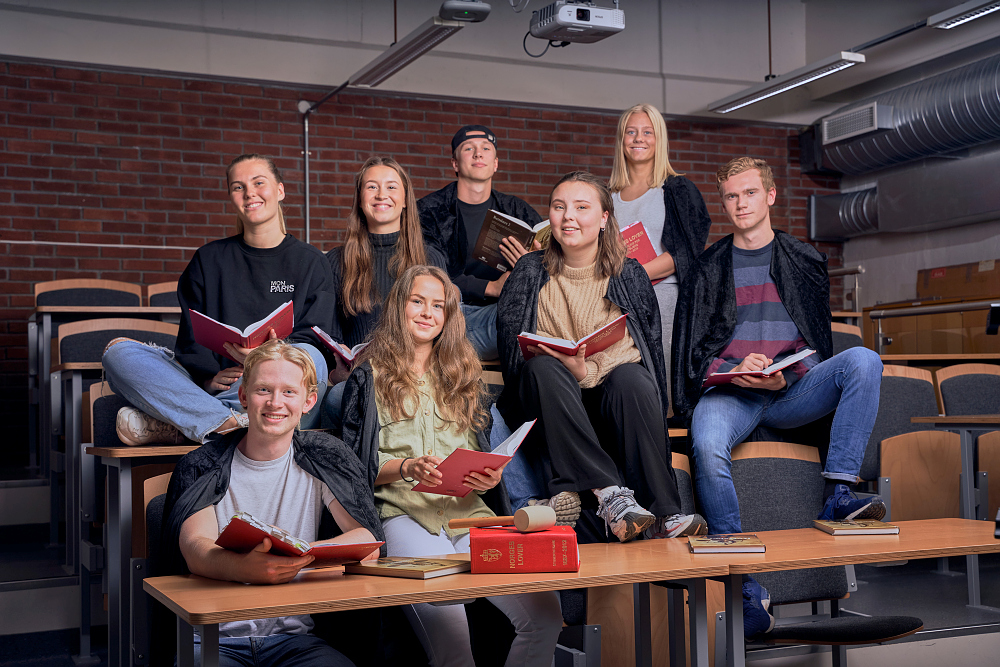 8 elever sitter i et auditorium med røde bøker i hendene og ser mot fotografen