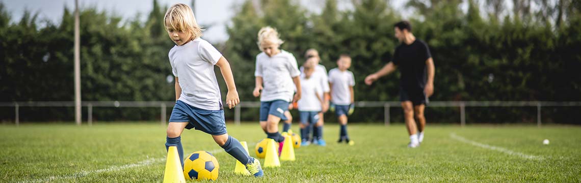 Barn i blå og hvite fotballdrakter fører ball mellom kjegler mens en voksen gir instruksjoner.