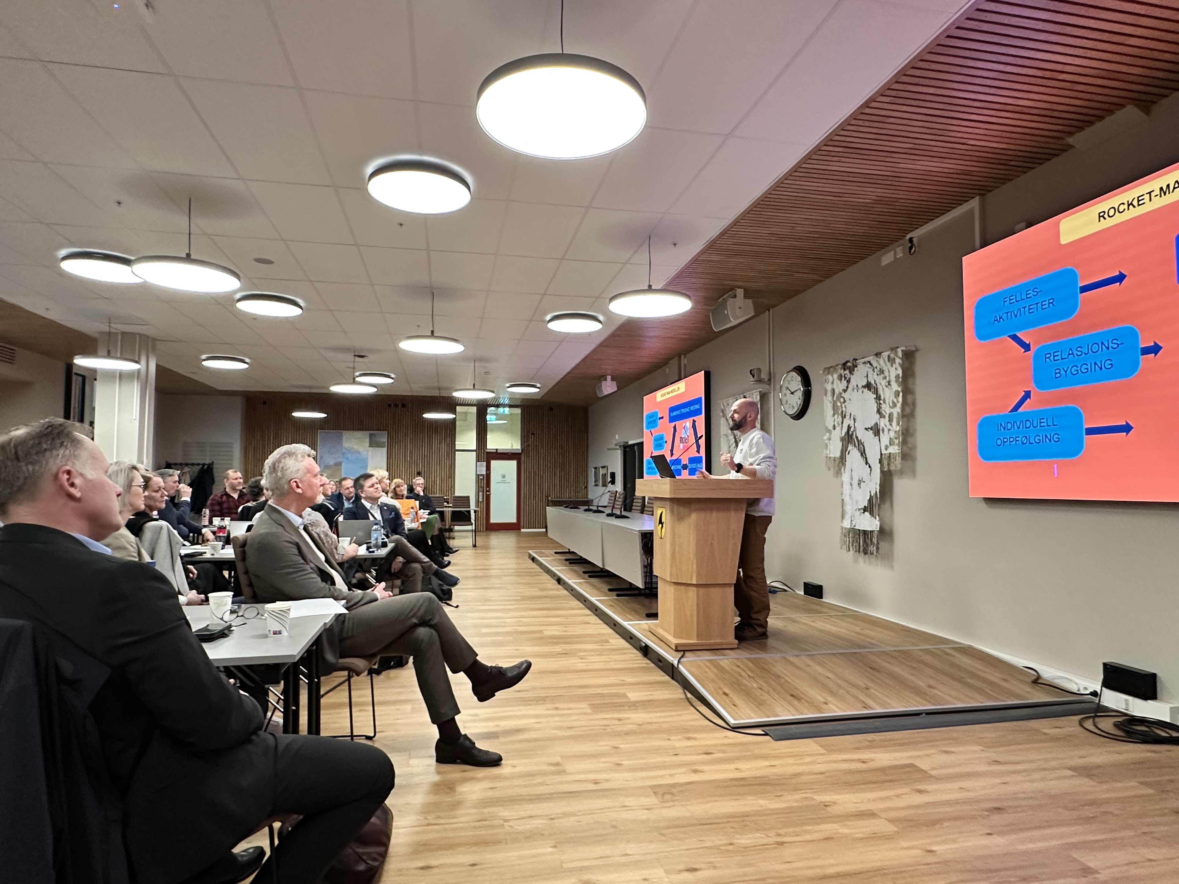 Bilde av en sal med mange deltagere som følger med på en som holder en presentasjon